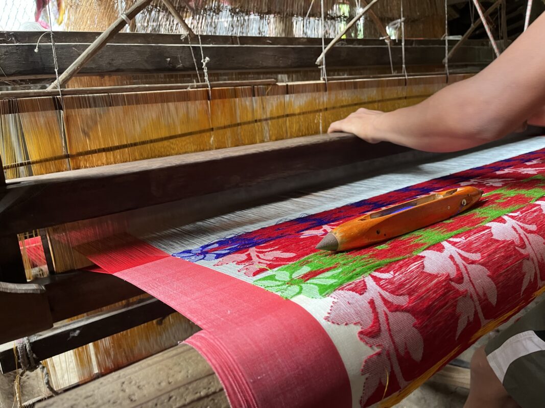Close-up of a Jamdani saree being handwoven on a traditional loom, showing intricate geometric and floral patterns in fine threads, with a weaver’s hands skillfully interlacing the delicate fabric.