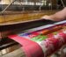 Close-up of a Jamdani saree being handwoven on a traditional loom, showing intricate geometric and floral patterns in fine threads, with a weaver’s hands skillfully interlacing the delicate fabric.