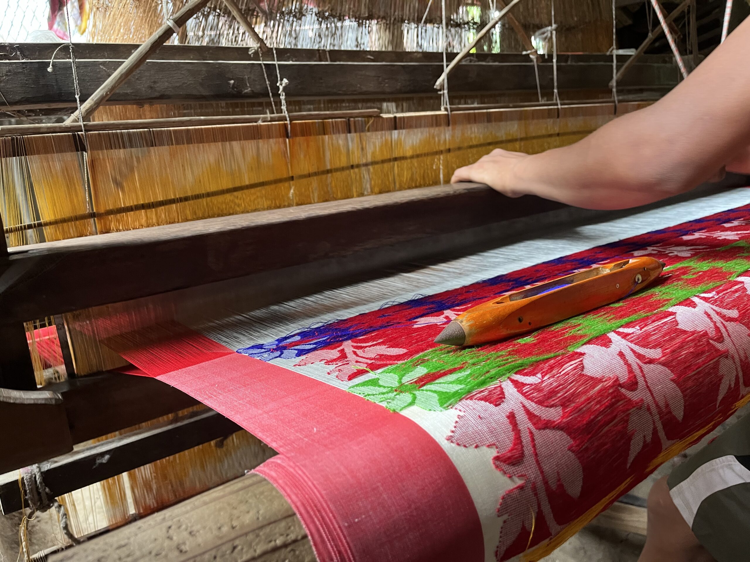Close-up of a Jamdani saree being handwoven on a traditional loom, showing intricate geometric and floral patterns in fine threads, with a weaver’s hands skillfully interlacing the delicate fabric.
